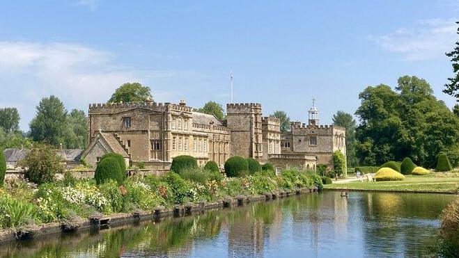 Forde Abbey as it looks today - a grand stone mansion with crenelated parapets stands next to a large rectangular lake. The building and herbaceous flower borders are reflected in the water.
