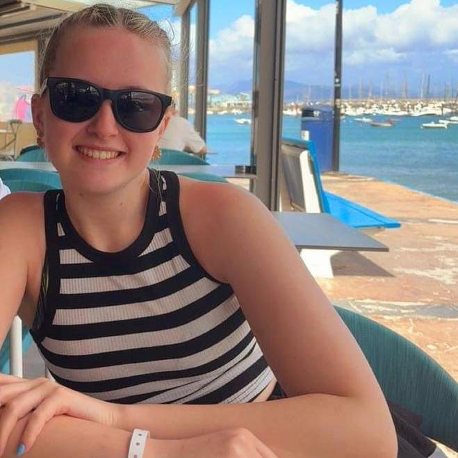 Meg with tied-back blonde hair, wearing sunglasses and a black and white striped vest top. She is sat down, smiling at the camera, with a marina behind her with blue water and white boats.