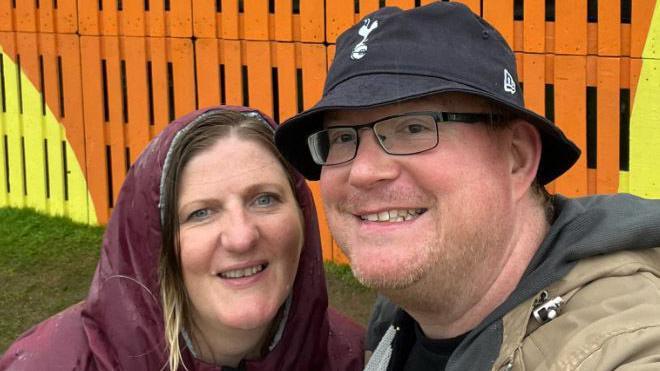 Kate and Colin Broadbent are smiling at the camera in front of orange and yellow wood panels. Colin is wearing a black bucket hat and black glasses, and Kate has brown hair, and is wearing a burgundy raincoat with the hood up