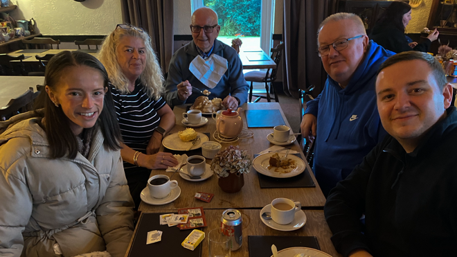 Hayley Morrison sits at a table in a restaurant with her family. There are cups of coffee in front of them and two plates. She wears a big puffa jacket.