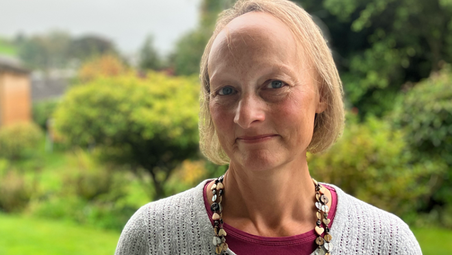 Jenny Tagney looks at the camera. She is wearing a wooden necklace, red top and grey cardigan, with her garden seen in the background. She has blonde, bobbed hair.