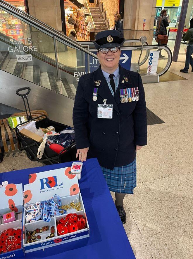 Tracy wearing military dress with multiple military medals stood in a shopping centre next to a Royal British Legion stall selling poppies. It is a full body shot and she is stood smiling at the camera. She is wearing glasses. 