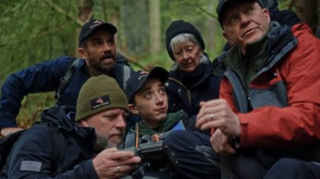Five people with Daniel in the middle crouched down in woodland looking around as if in search of something, with a man at the front holding a video camera