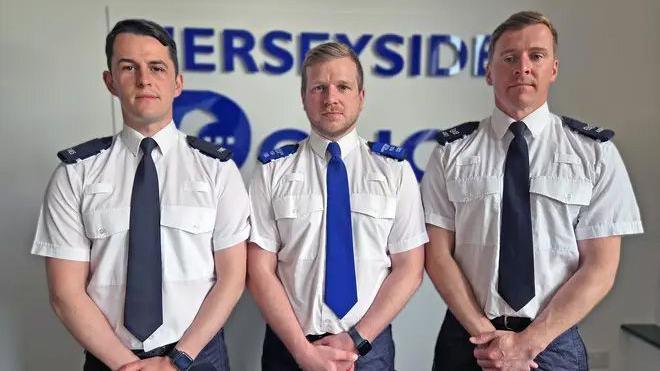 PC Luke Holden, PCSO Timothy Parry and Sgt Gregory Gillespie are standing in row in front of the Merseyside Police Federation logo. They are all wearing white short sleeved shirts with ties and have their hands clasped together in front of them.