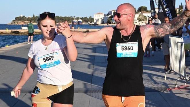 A girl with blonde/brown hair is running on prosthetic legs along the street holding a man's hand. She is wearing a white T-shirt with a race number pinned to it, black shorts and sunglasses on her head. The man has a shaved head and is wearing sunglasses, a necklace, a black vest with a race number pinned to it and orange shorts. They are both cheering and smiling