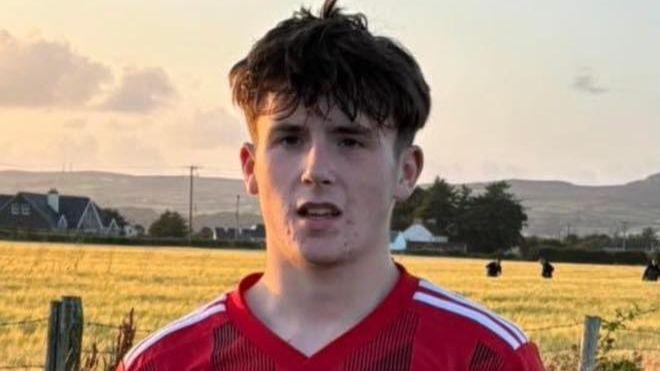 Tiernan Doherty-Kelly dressed in the red kit of Carndonagh FC, stands with his hands behind his back. He is standing on a football pitch, with a field of corn in the background 