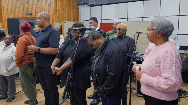 Men and women stand by microphones in the studio. They are wearing tops and trousers and some have coats on and are wearing glasses.