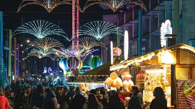 A Christmas market with lots of people lining a street with brightly lit stalls on the right side and multi-coloured lights lining the street