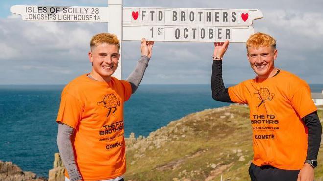 Two men in orange t-shirts standing in front of the sign for Land's End, on a cliff top overlooking the sea. The t-shirts are printed with a logo of a running brain, with The FTD Brothers written underneath. The Land's End sign has been customised to say "FTD Brothers, 1st October".