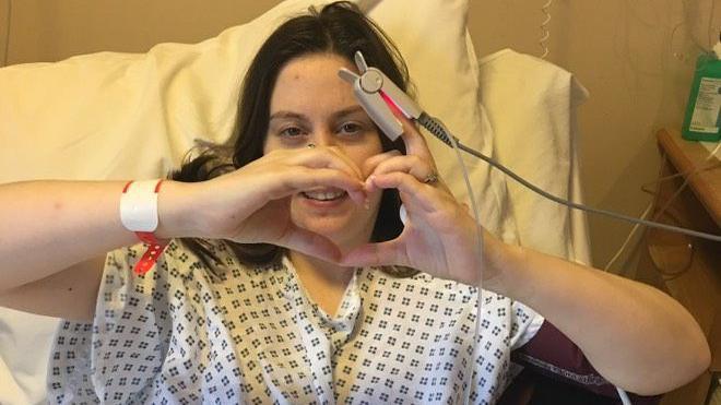 A woman in a hospital bed making a love heart shape with her hands in front of her face, with hospital clothes and wires attached to her