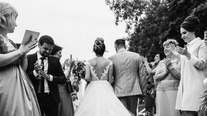 A black and white wedding picture, showing the back of a bride walking down the aisle outdoors surrounded by guests - one on the left is a man in a suit.