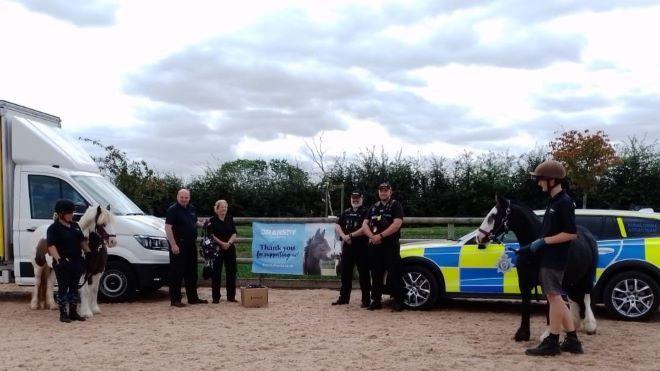 A demonstration of the collars in action, with two horses, police officers and members of the Bransby team standing in front of a police car. One of the horses has brown and white markings. The other has black and white markings 
