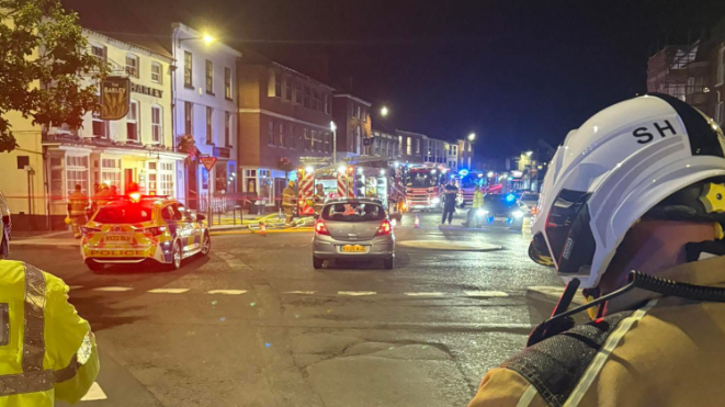 Multiple fire engines and a police car are pulled up outside a row of shops and businesses, with lights flashing. There is the back of a firefighter in the foreground