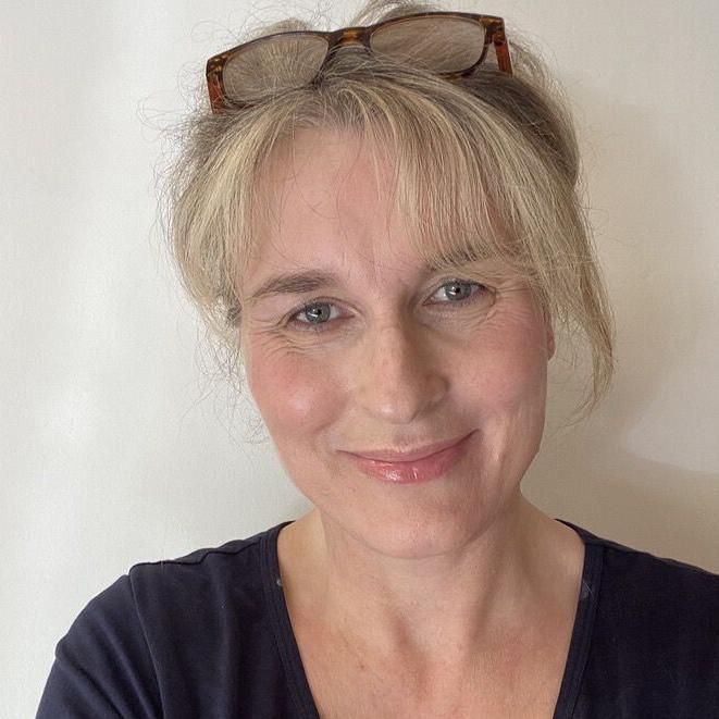 A selfie of a woman with tied-black blonde hair, wearing a black t shirt and brown glasses on top of her head. She is smiling and looking at the camera, and is stook in front of a plain white background.