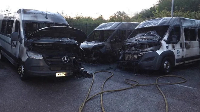 Three minibuses in a compound are completely blackened and burned out. A yellow fire service hose is on the ground near them.