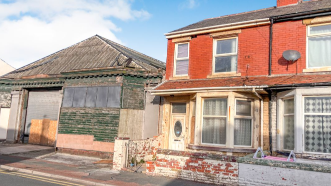 A wide shot of the front of the house. It has a white door, bay windows and orange brickwork. The front wall is discoloured, and the gate is open.