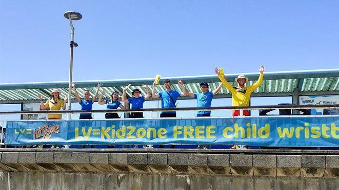 Seven people wearing blue and yellow t-shirts stand on a pier. They are waving bunches of bright yellow wristbands in the air.