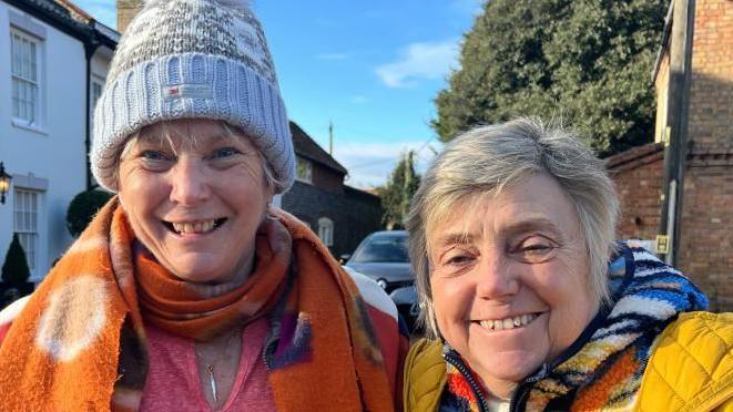 Two women standing by a road and smiling and looking straight at the camera. Caroline Giddings (left) is hearing a grey and blue hat, with a bobble on the top, a orange colourful scarf, and a pink top with a silver necklace round her neck. Denise Waters (right) has short grey hair, is hearing a colourful top with a yellow gilet. 