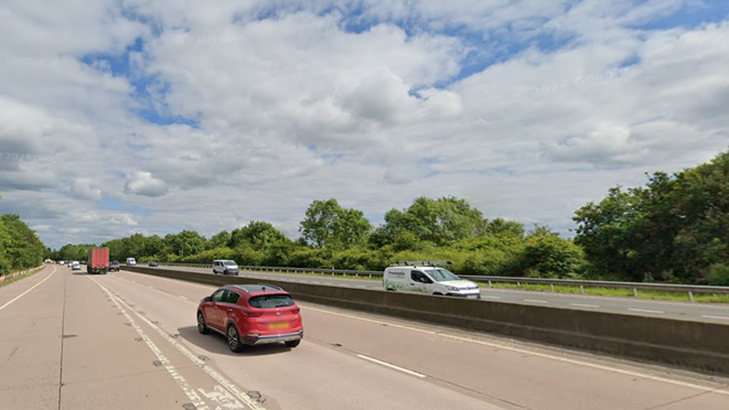 A motorway with a low concrete barrier in between the two carriageways. A red car is driving away from the shot and a white van towards it. The sky is blue in the background. 