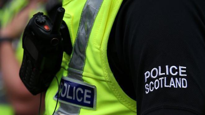 Police Scotland uniform, yellow bib with work police, and Police Scotland in bold white letters on a black sleeve.