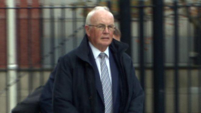 Frank Daly is an older man with short white hair. He is wearing a pair of rectangle glasses, a navy coat, a white shirt and a striped tie. The background of the courthouse is blurred.
