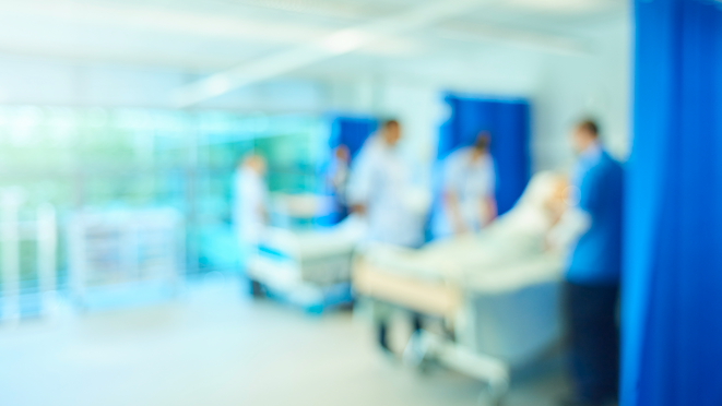 A senior female nursing sister demonstrates the the various equipment on the training ward whilst a male staff nurse shows the medical mannequin to another group of medical student nurses. They are all standing around the hospital beds. The whole scene is out of focus.