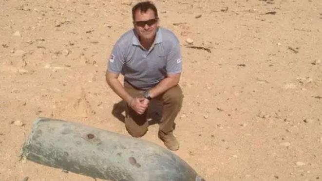 A man in a grey shirt and brown trousers kneeling down next to a large metal object in a dusty field.