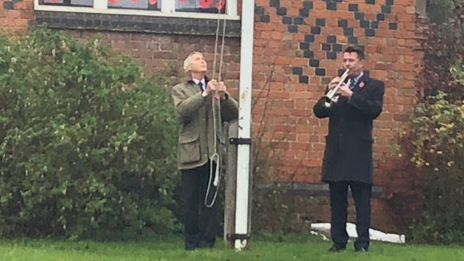 A man in a dark suit and coat, playing a bugle, alongside a man lowering a flag on a white flag pole. Behind them is a red brick building with darker bricks marking out a diamond pattern in the brick work with green bushes in front of the building