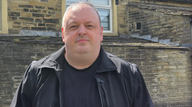 Man with cropped hair looking directly at the camera. He is wearing a black leather jacket and black shirt. He is standing in front of a house.