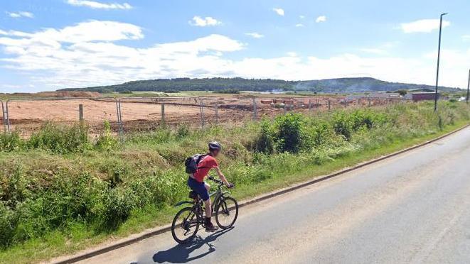 A cyclist on a road, with the beginning of a construction site to the left and low hills in the distance.