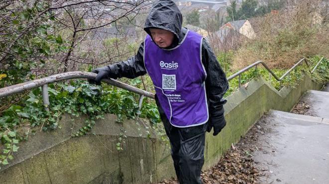 Derrick Downs climbing steep steps in very wet conditions wearing a mauve bib and looking soaked. There are houses down below in the distance