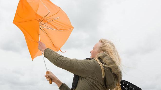 A woman in a brown raincoat struggles with an orange umbrella in strong winds and her bag is blowing behind her.