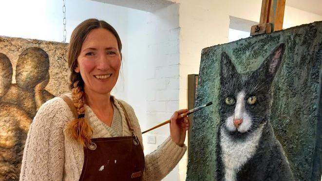 A woman with brown plaited hair, a beige knitted jumper and a maroon apron holding a brush next to a painting of a black and white cat on a green background.