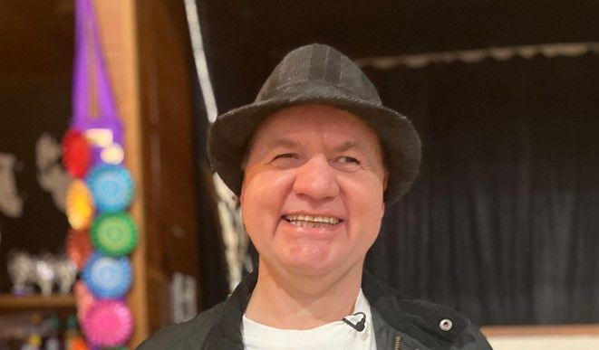 Blind dancer with trilby wearing a white T-shirt and black jacket in front of a black curtain in his dance studio.