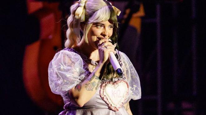 Melanie Martinez singing on stage with two-toned hair in braids, wearing a purple baby-doll dress