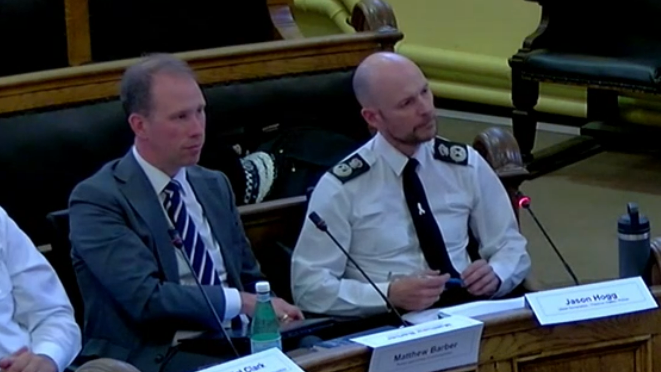 Jason Hogg, dressed in a white shirt and black tie, and holding a pen, sat next to Matthew Barber, in a dark blue suit and blue and white stripy tie. They are sat in Oxfordshire County Council's council chamber in County Hall in Oxford, which has yellow walls and benches with leather seating