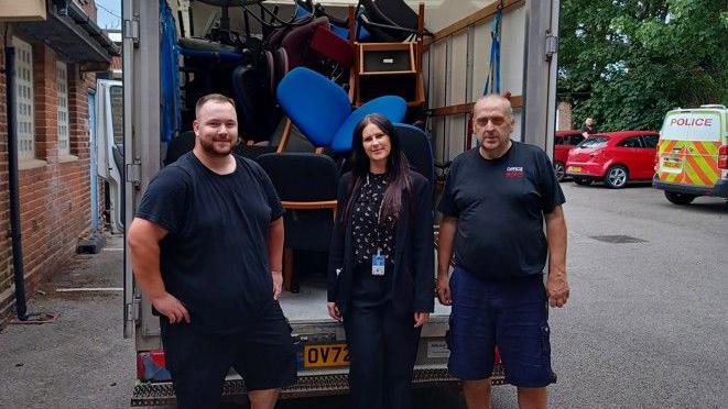 Two men stand either side of a woman at the back of a van filled with office chairs. The men are in navy t-shirts, shorts and work boots One, who as light brown hair with a beard, has his hands on his hips and is smiling. The other is balding and holds his hands by his side. The woman has long brown hair and is wearing a black suit and a black-and-white top and smiling. The three are standing in a car park with a police van in the background.