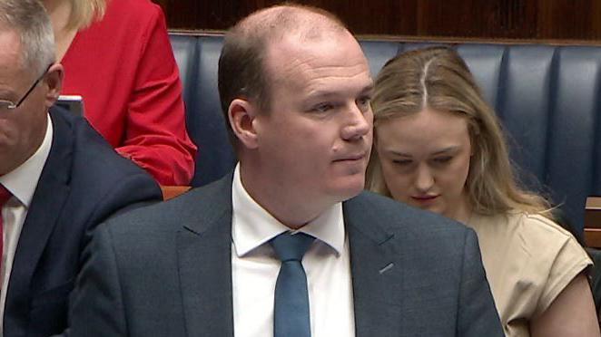 Gordon Lyons wearing navy suit and tie, speaking in the Assembly