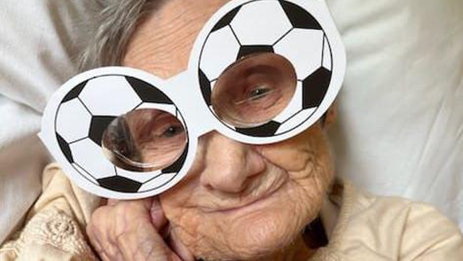 Irene Lane, an elderly woman with short grey hair, lies in a bed wearing comedy football-themed glasses.