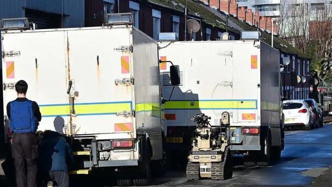 Two white police vans parked in a narrow street. There is a bomb disposal robot present