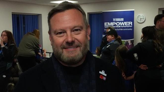 Mark Ashton stands inside a family hub and smiles at the camera. He has short dark and grey hair and beard. He wears a black coat. People can be seen talking behind him.