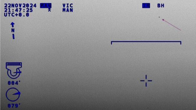 Infra-red camera image taken from a helicopter. There is an arrow pointing to an indistinct object. There are blue letters and numbers plus other types of on-screen information.