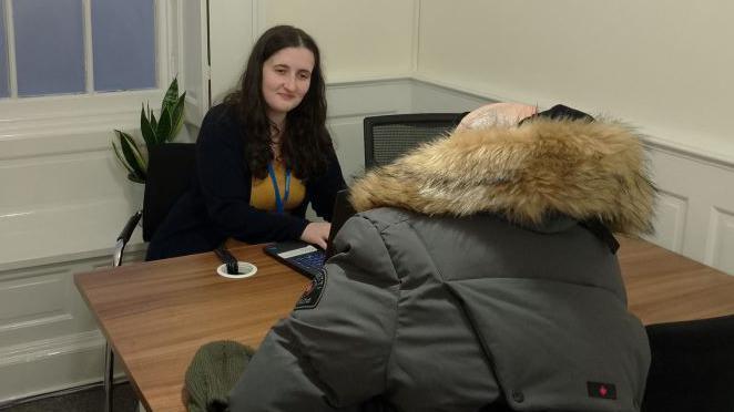 Two women sit on either side of a brown-topped desk, in a small room with cream walls in a small room, that has a window on the left. The woman in the foreground has her back to the camera and is wearing a parka-style coat, as she has a conversation with the other woman, who is wearing a security pass.
