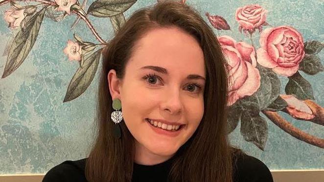 Sophie has shoulder-length brown hair and is wearing a black jumper and gold chain necklace. She is sitting in front of a wall with floral wallpaper.