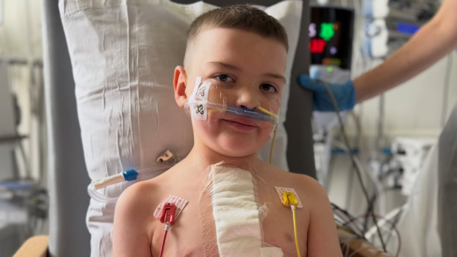 A young boy sitting in a hospital chair holding a football. He has a large bandage on his chest and several tubes and wires attached to his face and body.