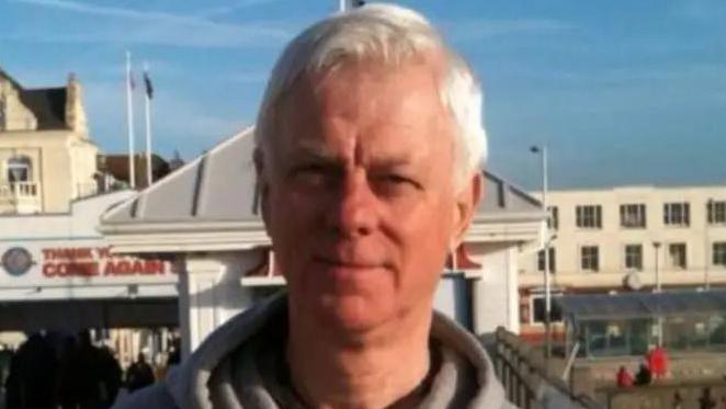 Paul Carter standing on a pier. He is wearing a grey hoodie. He has short, white hair. He is smiling and looking at the camera.