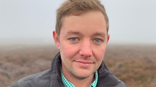 A man with blue eyes and blond hair in a grey coat standing in a blurred brown field