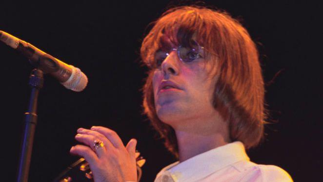 Liam with long hair holding a tambourine, wearing round glasses reminiscent of John Lennon and standing in front of a microphone