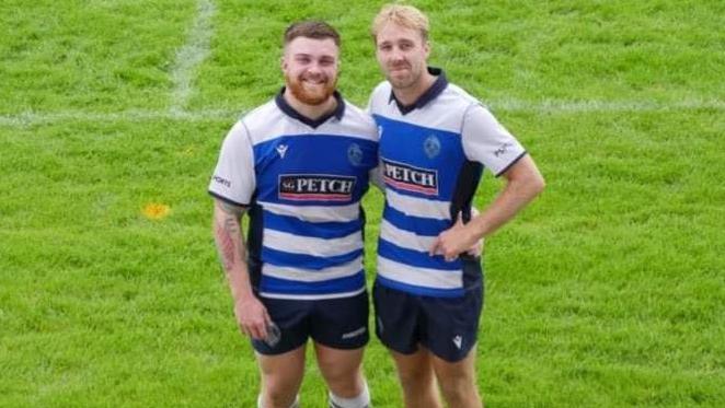 (L-R) Sam Clay and Josh Ree, who are both dressed in rugby kit, stand together on the pitch. Sam has a short beard and Josh has blond hair.