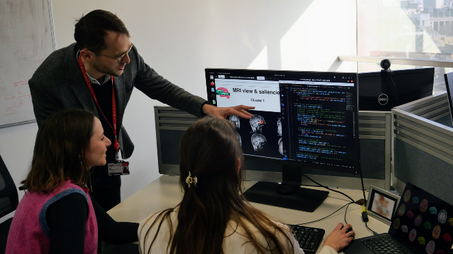 One male researcher stands, pointing at a computer screen, while two female researchers sit in front of the screen, with one holding a computer mouse.